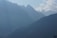 Two paragliders soar through a misty mountain landscape with rugged peaks and scattered clouds. The scene portrays a sense of adventure and tranquility against a backdrop of towering mountains.