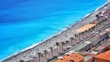A stunning beach scene with bright turquoise water and a wide sandy beach populated by numerous people enjoying the sun and sea. There is a promenade lined with palm trees and adjacent buildings with terracotta rooftops, adding to the vibrant coastal atmosphere.
