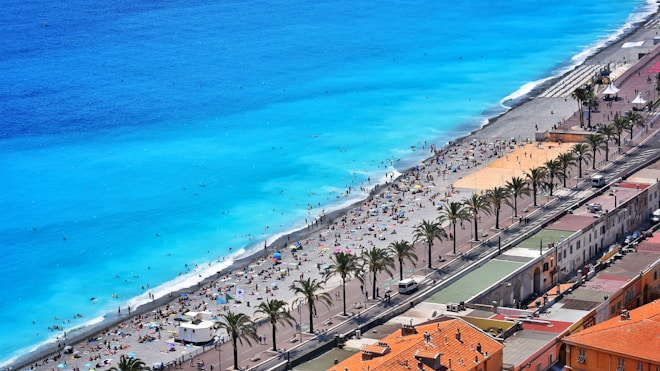A stunning beach scene with bright turquoise water and a wide sandy beach populated by numerous people enjoying the sun and sea. There is a promenade lined with palm trees and adjacent buildings with terracotta rooftops, adding to the vibrant coastal atmosphere.
