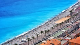 A stunning beach scene with bright turquoise water and a wide sandy beach populated by numerous people enjoying the sun and sea. There is a promenade lined with palm trees and adjacent buildings with terracotta rooftops, adding to the vibrant coastal atmosphere.