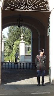 A uniformed guard stands at the entrance of a historic building with arched architecture. The interior showcases decorative wrought iron patterns above, and a paved path leads to a gated garden area with lush greenery in the background. Another guard is visible at a distance within the garden.