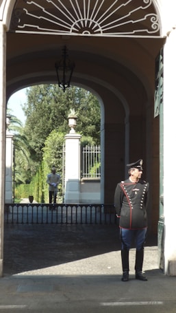 A uniformed guard stands at the entrance of a historic building with arched architecture. The interior showcases decorative wrought iron patterns above, and a paved path leads to a gated garden area with lush greenery in the background. Another guard is visible at a distance within the garden.