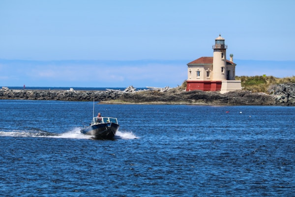 A small motorboat travels across a vast expanse of blue water, leaving a trail of white foam in its wake. In the background, a picturesque lighthouse with a red and beige facade stands on a rocky shoreline, surrounded by some vegetation. The sky is clear and blue, adding to the serene coastal scene.