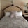 A neatly made bed with a tufted headboard featuring an ornate design. The bedding consists of a textured, striped comforter in a silver hue, with a matching decorative pillow in the center, flanked by plain white pillows. The wall behind the bed has a stucco finish, and there is a decorative wall sconce above the headboard. To the right, a wooden nightstand holds a traditional table lamp and a clock.