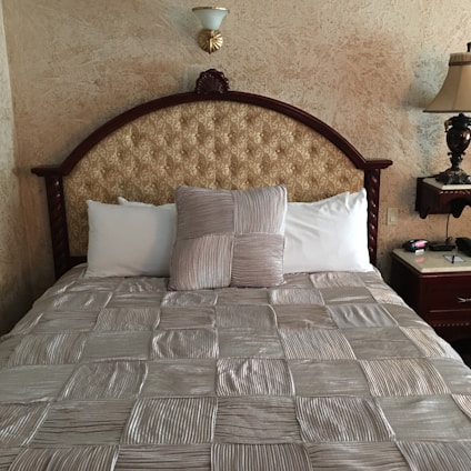 A neatly made bed with a tufted headboard featuring an ornate design. The bedding consists of a textured, striped comforter in a silver hue, with a matching decorative pillow in the center, flanked by plain white pillows. The wall behind the bed has a stucco finish, and there is a decorative wall sconce above the headboard. To the right, a wooden nightstand holds a traditional table lamp and a clock.
