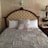 A neatly made bed with a tufted headboard featuring an ornate design. The bedding consists of a textured, striped comforter in a silver hue, with a matching decorative pillow in the center, flanked by plain white pillows. The wall behind the bed has a stucco finish, and there is a decorative wall sconce above the headboard. To the right, a wooden nightstand holds a traditional table lamp and a clock.