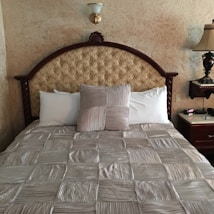 A neatly made bed with a tufted headboard featuring an ornate design. The bedding consists of a textured, striped comforter in a silver hue, with a matching decorative pillow in the center, flanked by plain white pillows. The wall behind the bed has a stucco finish, and there is a decorative wall sconce above the headboard. To the right, a wooden nightstand holds a traditional table lamp and a clock.