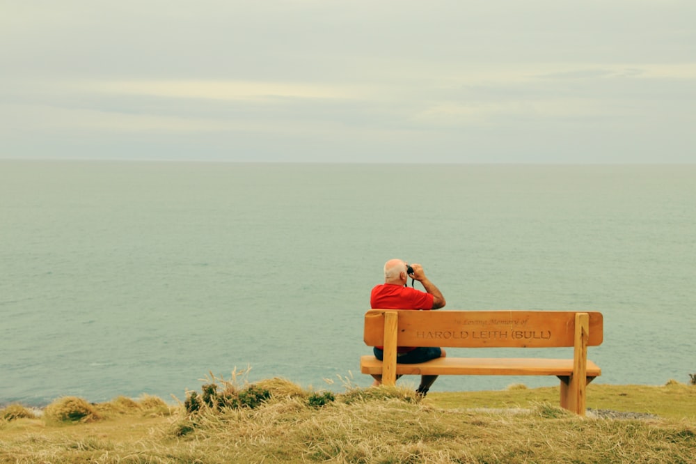 man wearing red t-shirt sitting on brown wooden bench facing on body of water