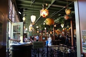 A cozy restaurant interior with various hanging decorative lamps illuminating the space. The setting includes wooden furniture, green walls, and potted plants, creating a warm and inviting atmosphere. Diners are seated at tables, and staff are seen moving around.