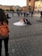 A couple in wedding attire sits on the ground in a city square, surrounded by a crowd of people. The bride is wearing a white gown and the groom is in a dark suit. There is a photographer with an orange backpack taking pictures of them. Historic buildings and a statue are visible in the background.