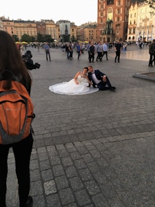 A couple in wedding attire sits on the ground in a city square, surrounded by a crowd of people. The bride is wearing a white gown and the groom is in a dark suit. There is a photographer with an orange backpack taking pictures of them. Historic buildings and a statue are visible in the background.