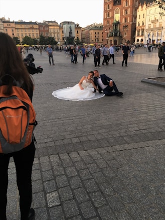 A couple in wedding attire sits on the ground in a city square, surrounded by a crowd of people. The bride is wearing a white gown and the groom is in a dark suit. There is a photographer with an orange backpack taking pictures of them. Historic buildings and a statue are visible in the background.