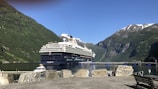 A red cruise ship anchored at a quiet, picturesque harbor with mountains in the background.