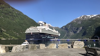A large cruise ship sits in a serene harbor surrounded by majestic mountains with patches of snow at their peaks. The water is calm, reflecting the clear blue sky above. In the foreground, large rocks and a small dock are visible.
