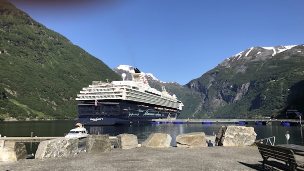 A large cruise ship sits in a serene harbor surrounded by majestic mountains with patches of snow at their peaks. The water is calm, reflecting the clear blue sky above. In the foreground, large rocks and a small dock are visible.