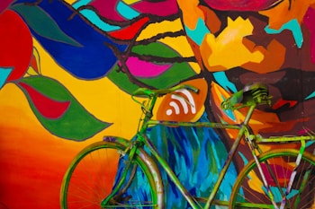 A colorful mural featuring abstract patterns and shapes in vivid hues covers a wall. In the foreground, a green bicycle is parked against this vibrant backdrop. The mural includes various outlines of leaves and abstract designs in red, blue, yellow, orange, pink, and green.