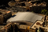 An aerial view of a cityscape featuring a large modern building with a distinctive curved roof made of glass or solar panels. The structure stands out among the surrounding traditional buildings with flat roofs and varied architectural designs. The play of light and shadow creates an interesting pattern on the roof.