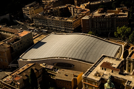 An aerial view of a cityscape featuring a large modern building with a distinctive curved roof made of glass or solar panels. The structure stands out among the surrounding traditional buildings with flat roofs and varied architectural designs. The play of light and shadow creates an interesting pattern on the roof.