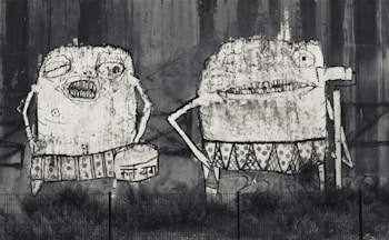 Two cartoonish figures are painted on a large wall. They have exaggerated features such as wide mouths and large eyes. The figures appear to be humanoid with intricate patterns on their clothing. The wall is weathered, contributing to an overall gritty appearance.