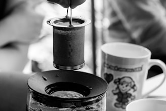 A tea infuser is held above a glass teapot, with water visibly inside the pot. Nearby, there are two mugs with unique designs; one features an illustration of a child and a heart. The setting is likely a cozy, relaxed environment.