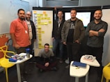 A group of six men are gathered in a room, standing and sitting around a set of whiteboards, which are covered with sticky notes. They appear to be engaged in a collaborative or planning session. There are tables with stationery supplies, notebooks, and a blue water bottle. The setting suggests a casual but focused work environment.