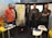 A group of six men are gathered in a room, standing and sitting around a set of whiteboards, which are covered with sticky notes. They appear to be engaged in a collaborative or planning session. There are tables with stationery supplies, notebooks, and a blue water bottle. The setting suggests a casual but focused work environment.
