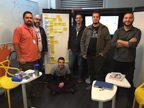 A group of six men are gathered in a room, standing and sitting around a set of whiteboards, which are covered with sticky notes. They appear to be engaged in a collaborative or planning session. There are tables with stationery supplies, notebooks, and a blue water bottle. The setting suggests a casual but focused work environment.