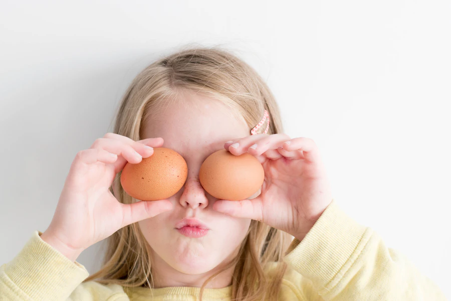 Playful girl making silly faces with eggs at the kitchen table
