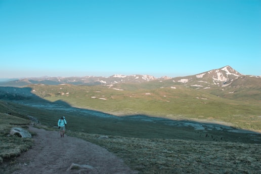 A lone hiker walks along a dirt path with trekking poles, surrounded by a vast, open landscape. Rolling green hills stretch into the distance, with patches of snow on a prominent mountain range under a clear blue sky.