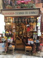 A small, colorful shop filled with a variety of products including hanging sausages, baskets of fruit, and vibrant packages. The exterior is adorned with plants and signs, giving it a rustic and inviting appearance. The interior is densely packed with diverse items for sale.