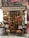 A small, colorful shop filled with a variety of products including hanging sausages, baskets of fruit, and vibrant packages. The exterior is adorned with plants and signs, giving it a rustic and inviting appearance. The interior is densely packed with diverse items for sale.