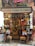 A small, colorful shop filled with a variety of products including hanging sausages, baskets of fruit, and vibrant packages. The exterior is adorned with plants and signs, giving it a rustic and inviting appearance. The interior is densely packed with diverse items for sale.