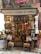 A small, colorful shop filled with a variety of products including hanging sausages, baskets of fruit, and vibrant packages. The exterior is adorned with plants and signs, giving it a rustic and inviting appearance. The interior is densely packed with diverse items for sale.