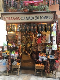 A small, colorful shop filled with a variety of products including hanging sausages, baskets of fruit, and vibrant packages. The exterior is adorned with plants and signs, giving it a rustic and inviting appearance. The interior is densely packed with diverse items for sale.