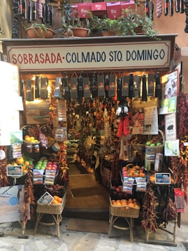 A small, colorful shop filled with a variety of products including hanging sausages, baskets of fruit, and vibrant packages. The exterior is adorned with plants and signs, giving it a rustic and inviting appearance. The interior is densely packed with diverse items for sale.