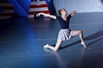 A dancer performs a dramatic pose on stage, featuring an athletic stance with one knee on the ground and the other leg extended. The dancer wears a dark top and a floral skirt, captured in mid-performance under bright stage lights. American flag-themed decorations are visible in the background.