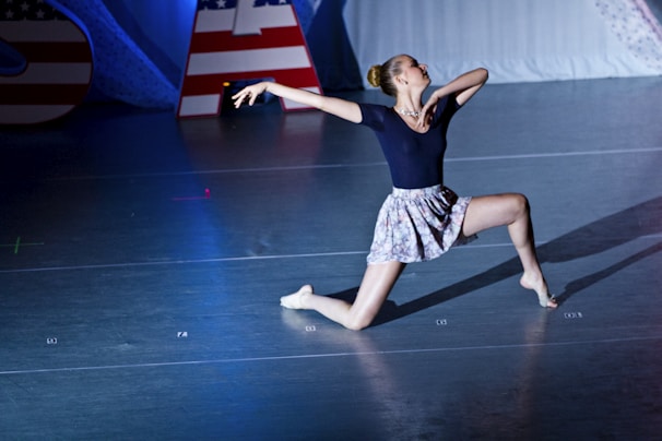 A dancer performs a dramatic pose on stage, featuring an athletic stance with one knee on the ground and the other leg extended. The dancer wears a dark top and a floral skirt, captured in mid-performance under bright stage lights. American flag-themed decorations are visible in the background.