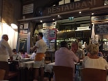 A bustling restaurant scene with a warm, inviting atmosphere. Several people are seated at tables covered with white tablecloths, engaged in conversation and dining. A waiter is attentively serving the guests. The interior is decorated with a variety of signage, chalkboards with menus, and bottles displayed on shelves, contributing to a cozy and lively ambiance.
