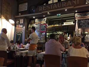 A bustling restaurant scene with a warm, inviting atmosphere. Several people are seated at tables covered with white tablecloths, engaged in conversation and dining. A waiter is attentively serving the guests. The interior is decorated with a variety of signage, chalkboards with menus, and bottles displayed on shelves, contributing to a cozy and lively ambiance.