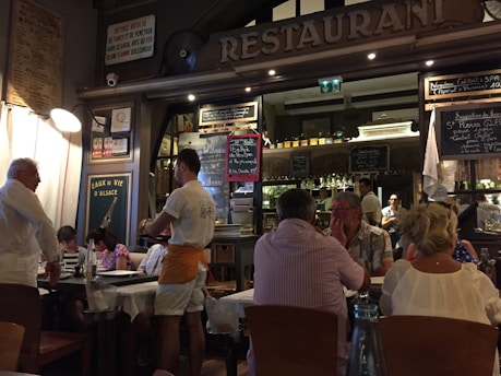 A bustling restaurant scene with a warm, inviting atmosphere. Several people are seated at tables covered with white tablecloths, engaged in conversation and dining. A waiter is attentively serving the guests. The interior is decorated with a variety of signage, chalkboards with menus, and bottles displayed on shelves, contributing to a cozy and lively ambiance.