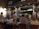 A bustling restaurant scene with a warm, inviting atmosphere. Several people are seated at tables covered with white tablecloths, engaged in conversation and dining. A waiter is attentively serving the guests. The interior is decorated with a variety of signage, chalkboards with menus, and bottles displayed on shelves, contributing to a cozy and lively ambiance.