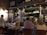 A bustling restaurant scene with a warm, inviting atmosphere. Several people are seated at tables covered with white tablecloths, engaged in conversation and dining. A waiter is attentively serving the guests. The interior is decorated with a variety of signage, chalkboards with menus, and bottles displayed on shelves, contributing to a cozy and lively ambiance.