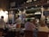 A bustling restaurant scene with a warm, inviting atmosphere. Several people are seated at tables covered with white tablecloths, engaged in conversation and dining. A waiter is attentively serving the guests. The interior is decorated with a variety of signage, chalkboards with menus, and bottles displayed on shelves, contributing to a cozy and lively ambiance.