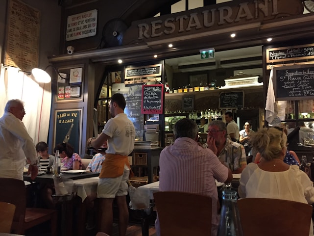 A bustling restaurant scene with a warm, inviting atmosphere. Several people are seated at tables covered with white tablecloths, engaged in conversation and dining. A waiter is attentively serving the guests. The interior is decorated with a variety of signage, chalkboards with menus, and bottles displayed on shelves, contributing to a cozy and lively ambiance.