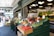 A bustling market stall displays a vibrant array of fresh produce, including tomatoes, asparagus, bell peppers, avocados, and pineapples. Arranged neatly in wooden crates, the fruits and vegetables are accompanied by price tags. The market area is lively with people walking by, some examining the goods. Overhead lights illuminate the colorful assortment, and a sign above reads 'Corner Produce'. In the background, additional greenery and vegetables are visible on shelves.