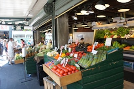 A bustling market stall displays a vibrant array of fresh produce, including tomatoes, asparagus, bell peppers, avocados, and pineapples. Arranged neatly in wooden crates, the fruits and vegetables are accompanied by price tags. The market area is lively with people walking by, some examining the goods. Overhead lights illuminate the colorful assortment, and a sign above reads 'Corner Produce'. In the background, additional greenery and vegetables are visible on shelves.