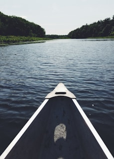 A canoe glides along a calm, expansive river, surrounded by lush green trees on both banks. The perspective from the canoe's bow emphasizes the open, serene water, which stretches into the distance under a clear sky.
