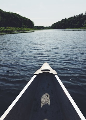 A canoe glides along a calm, expansive river, surrounded by lush green trees on both banks. The perspective from the canoe's bow emphasizes the open, serene water, which stretches into the distance under a clear sky.