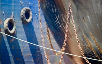 A close-up view of the hull of a ship displaying two large tires hanging on the side, likely used as fenders. Multiple heavy chains are draped across the side, casting shadows against the metal surface. The dark blue metallic hull shows some signs of rust and weathering, contributing to a rugged, industrial texture.