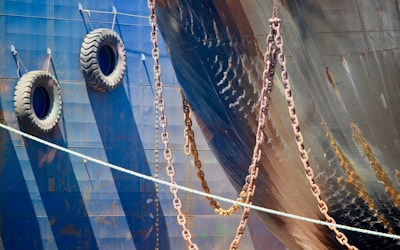 A close-up view of the hull of a ship displaying two large tires hanging on the side, likely used as fenders. Multiple heavy chains are draped across the side, casting shadows against the metal surface. The dark blue metallic hull shows some signs of rust and weathering, contributing to a rugged, industrial texture.